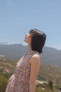 A woman in a floral dress faces the sun with mountains in the background.