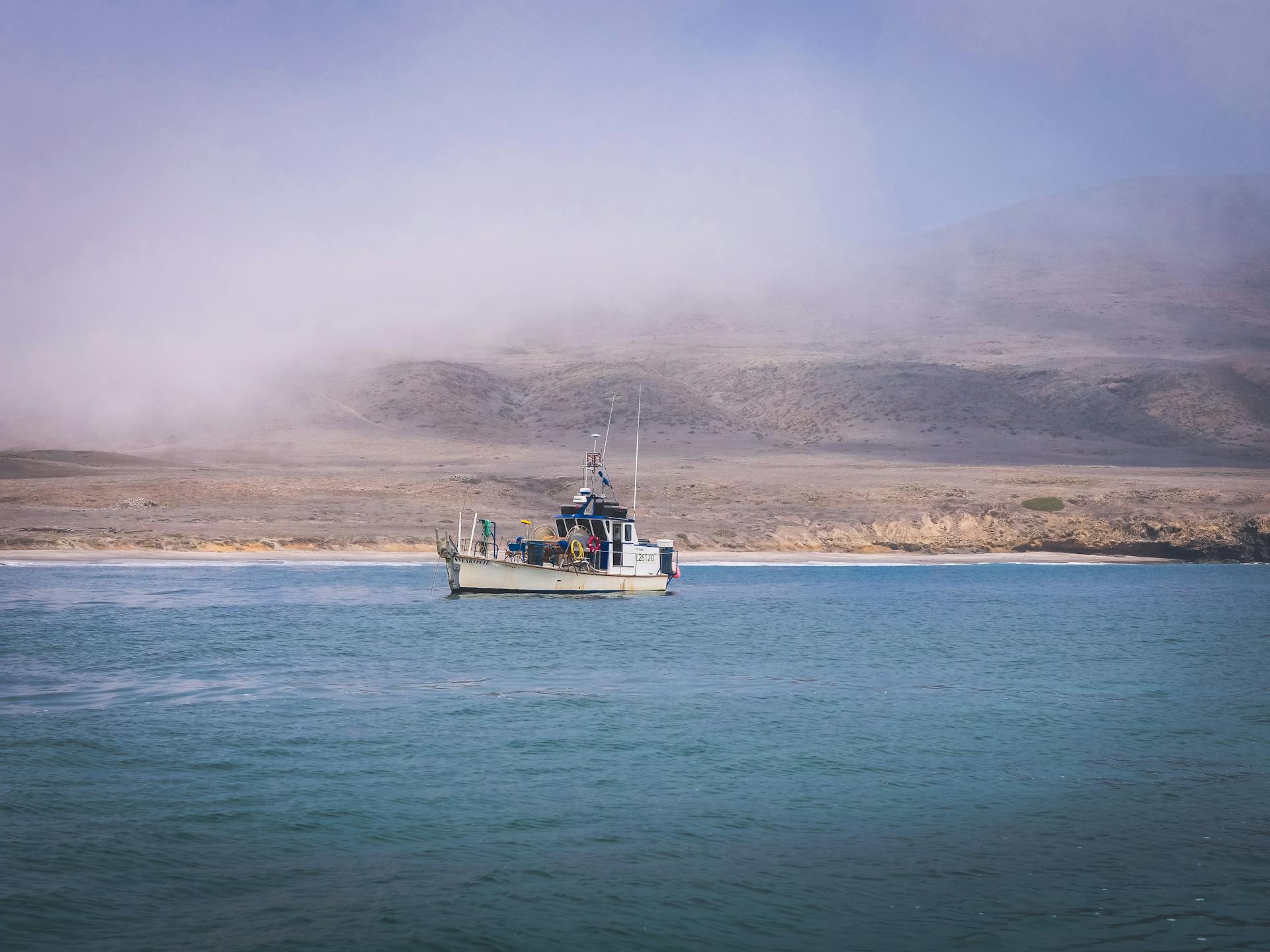 A fishing vessel navigates calm ocean waters under a misty sky, near a rugged coastline.