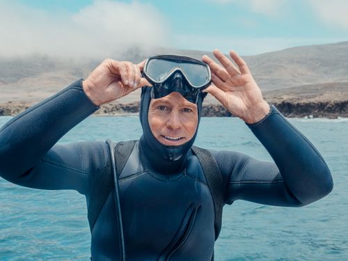 Free A smiling diver adjusts his goggles while wearing a wetsuit near a rocky coastline. Stock Photo