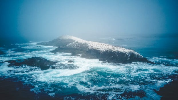 A stunning aerial capture of a rocky island in the ocean surrounded by crashing waves, under a misty sky.