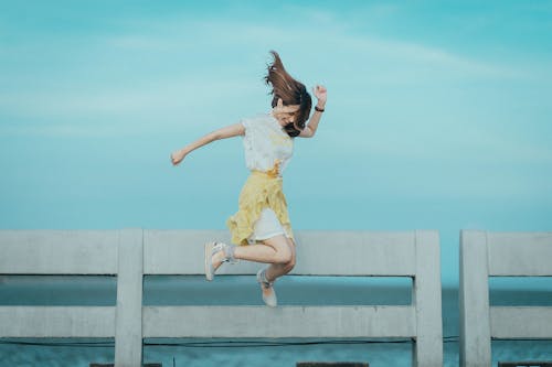 Free Jumpshot Photography of Woman in White and Yellow Dress Near Body of Water Stock Photo