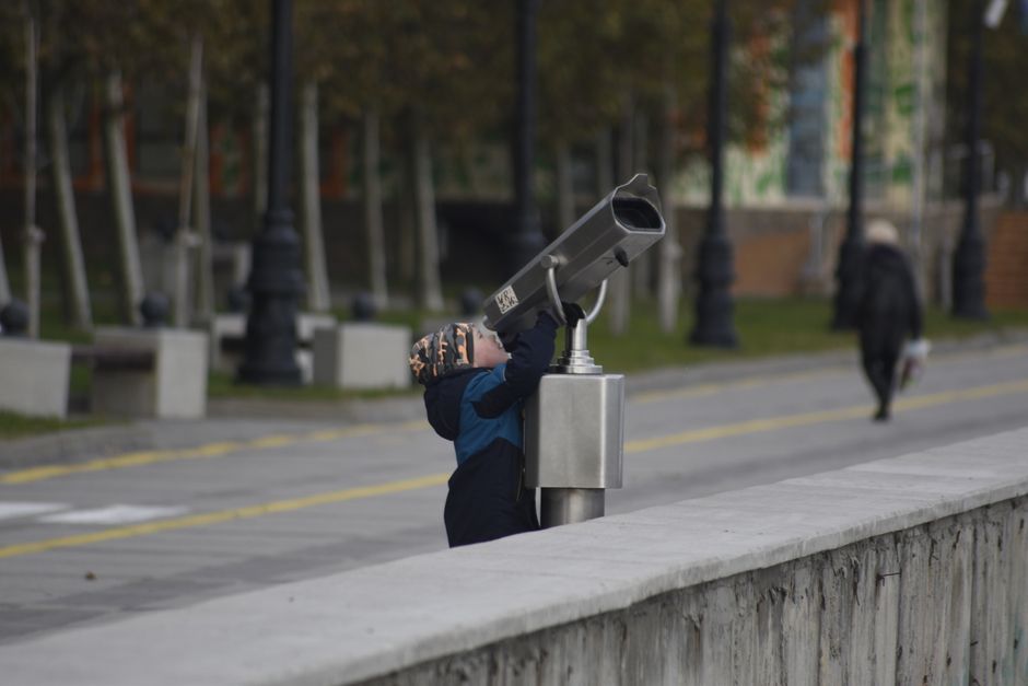 A child in winter clothes uses public binoculars in a park setting, showcasing curiosity and exploration.