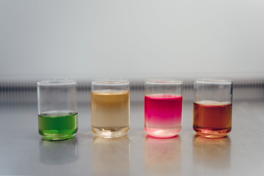 Free Four glasses filled with colorful liquids on a laboratory table, showcasing a chemical experiment. Stock Photo