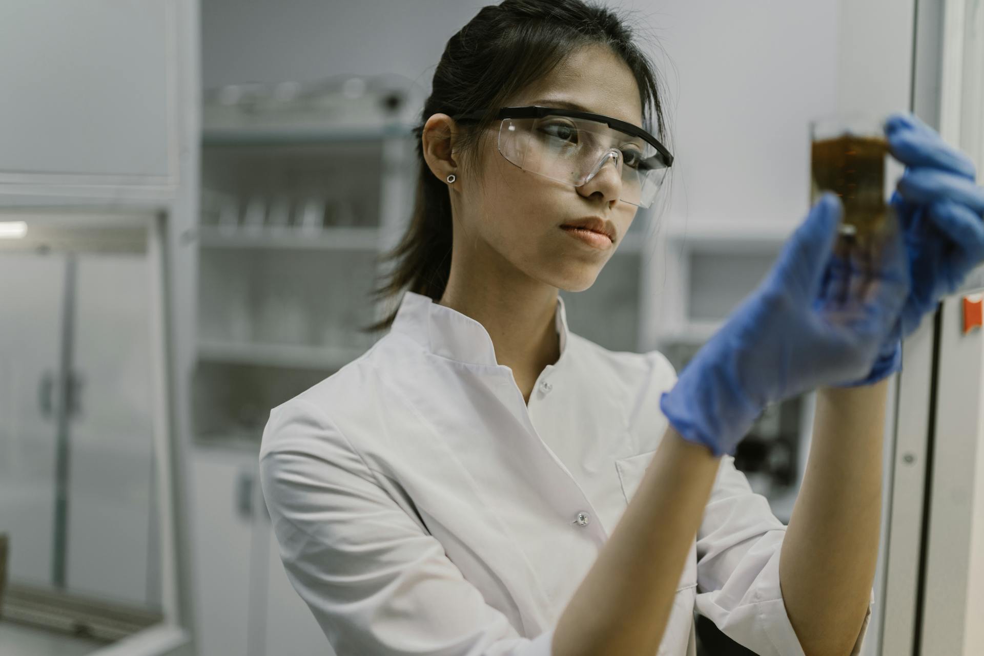 Young female scientist in lab coat examining a brown liquid in a glass beaker inside a laboratory.