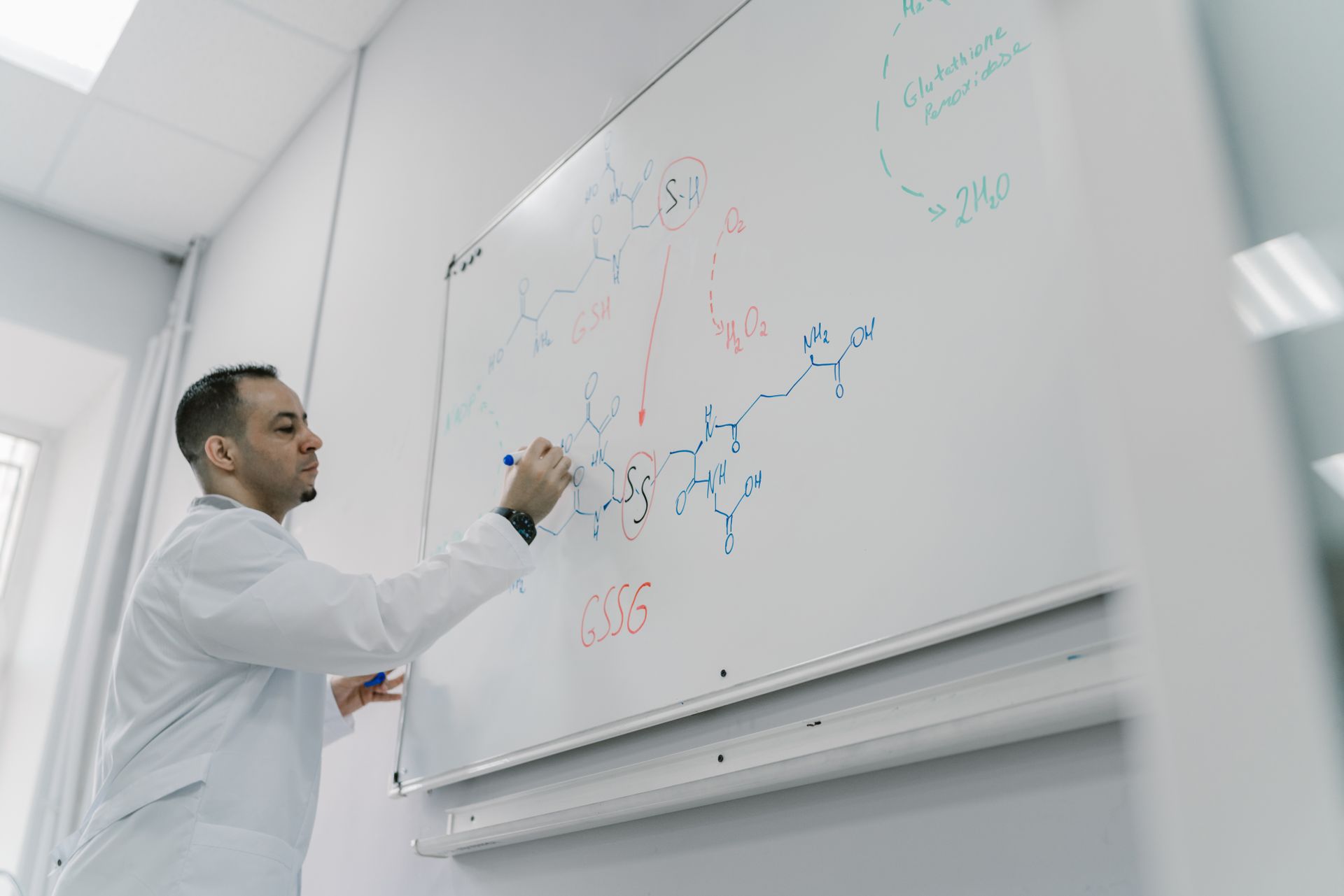 Male teacher explains chemical equation on whiteboard in science classroom setting.