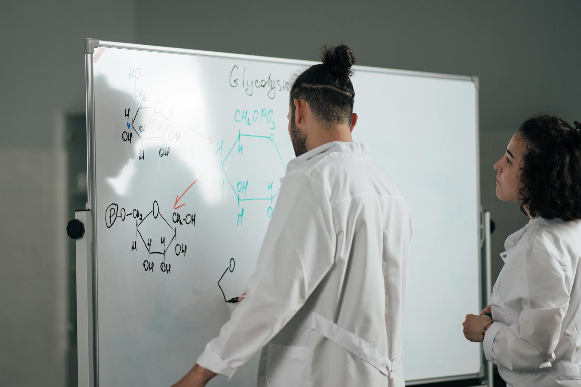 Two scientists in lab coats writing and discussing chemical formulas on a whiteboard in a laboratory setting.