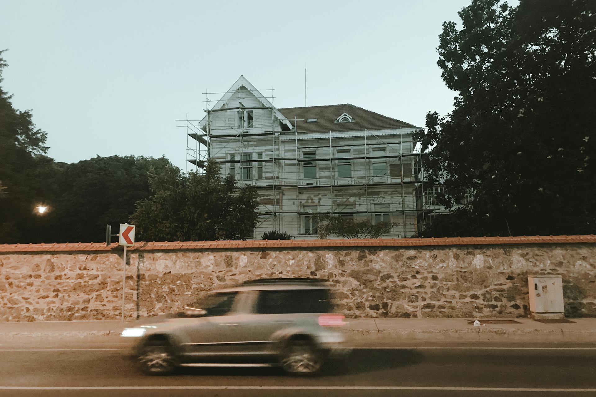 A blurred car drives past a historic house under renovation with scaffolding, set against a tranquil evening sky.