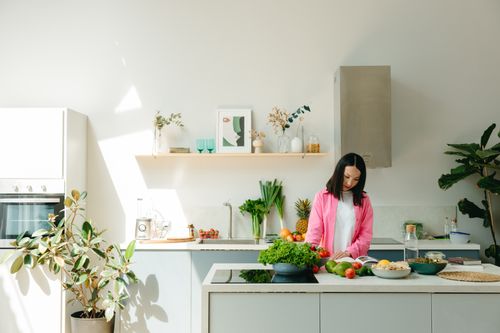 Free A woman in pink prepares fresh vegetables in a sunlit, modern kitchen, creating a vibrant cooking scene. Stock Photo