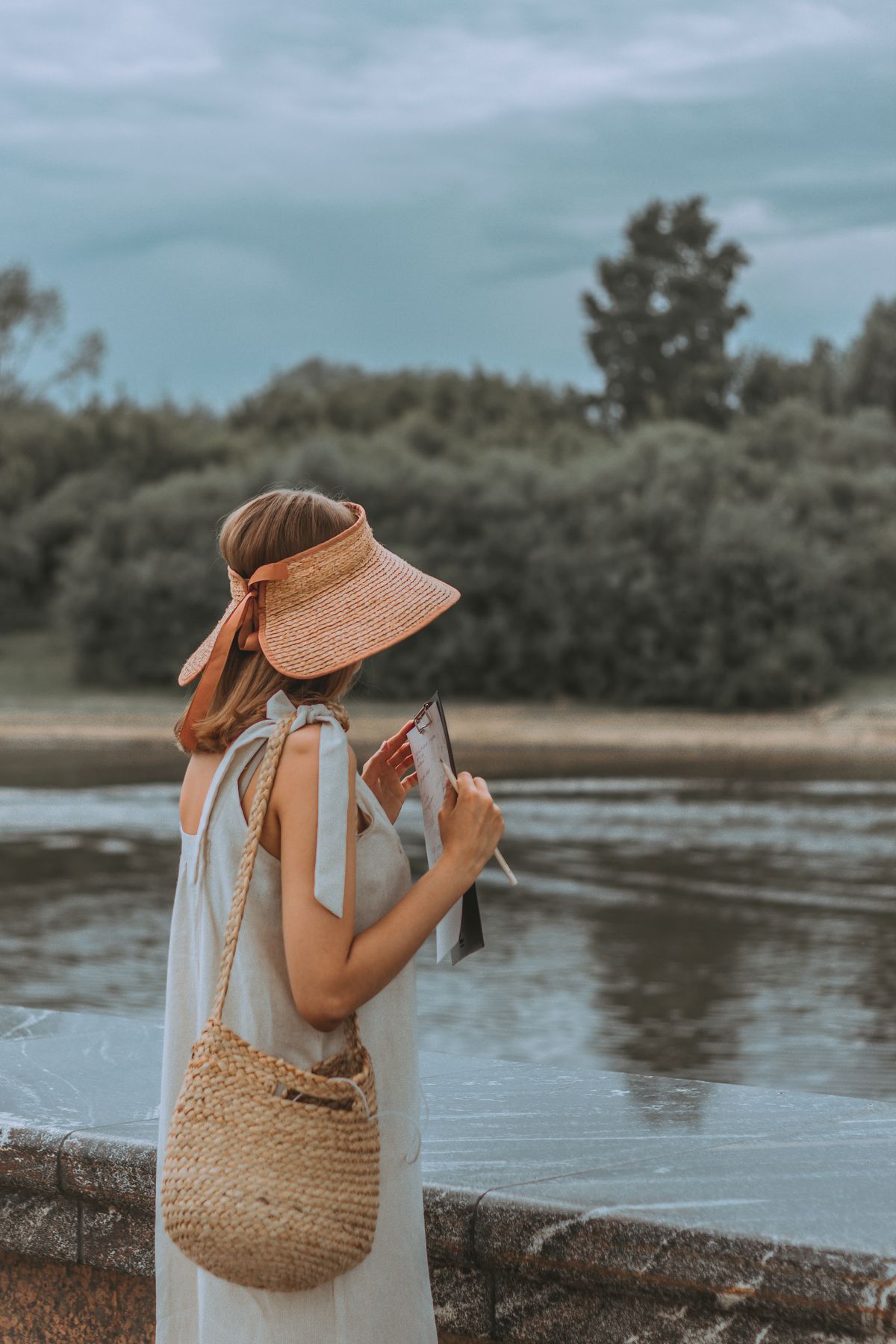 Woman in a sun visor holding a notebook beside a lake with a straw bag