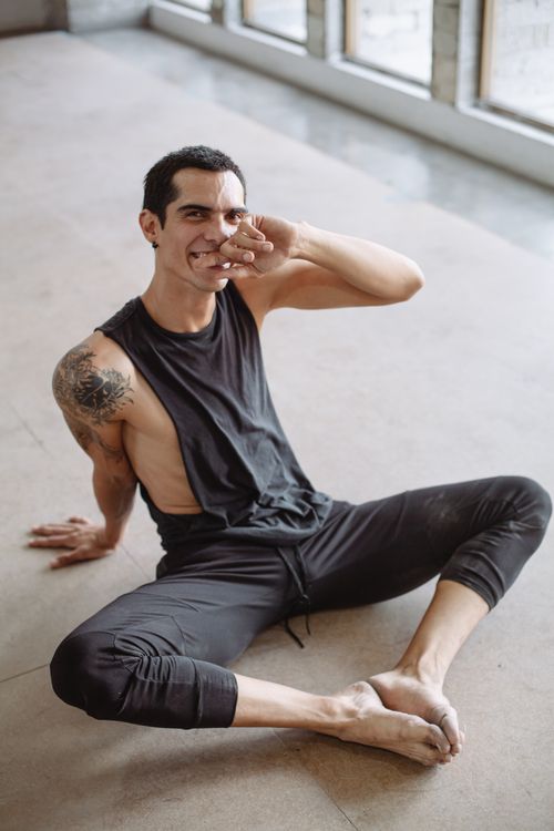 Free Man in casual black attire, smiling and sitting barefoot indoors with tattooed arm. Stock Photo