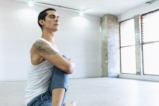 A ballet dancer in a white tank top sits in a dance studio, deep in thought.