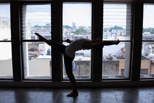 Backlit silhouette of a dancer performing a stretch in front of large urban windows, highlighting flexibility.