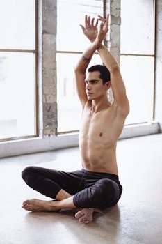 Tattooed man stretching while seated in a yoga pose inside a bright room.