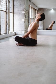 A shirtless male ballet dancer sits on the floor, stretching in a dance studio with a mirror reflection.