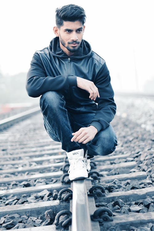 免费 黑色的皮革拉链帽衫和黑色牛仔牛仔裤的男人坐在沙哑的早晨包围着岩石的灰色金属火车栏杆上 素材图片