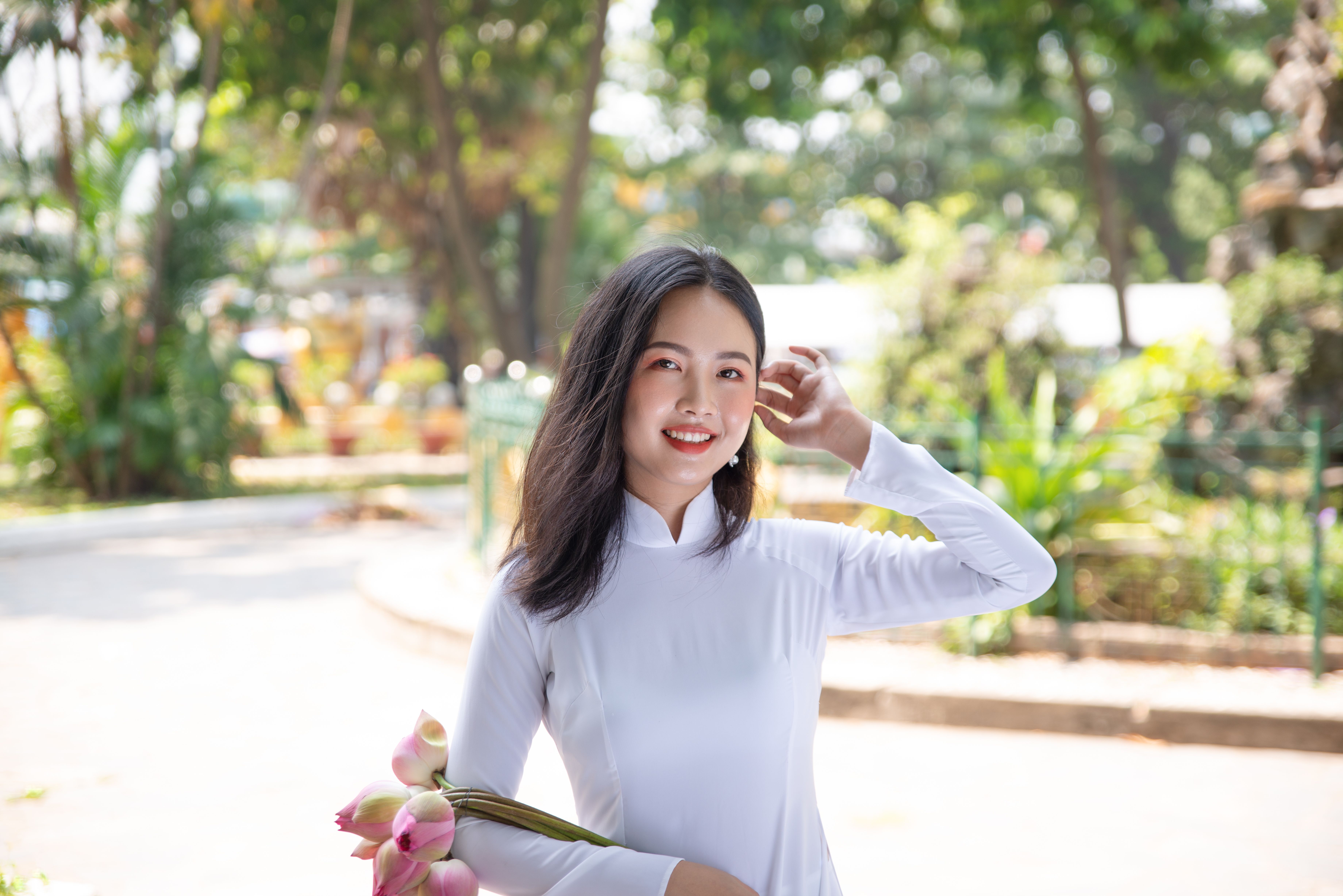 Asian woman in ao dai with lotus flowers, smiling outdoors. Captures cultural elegance.