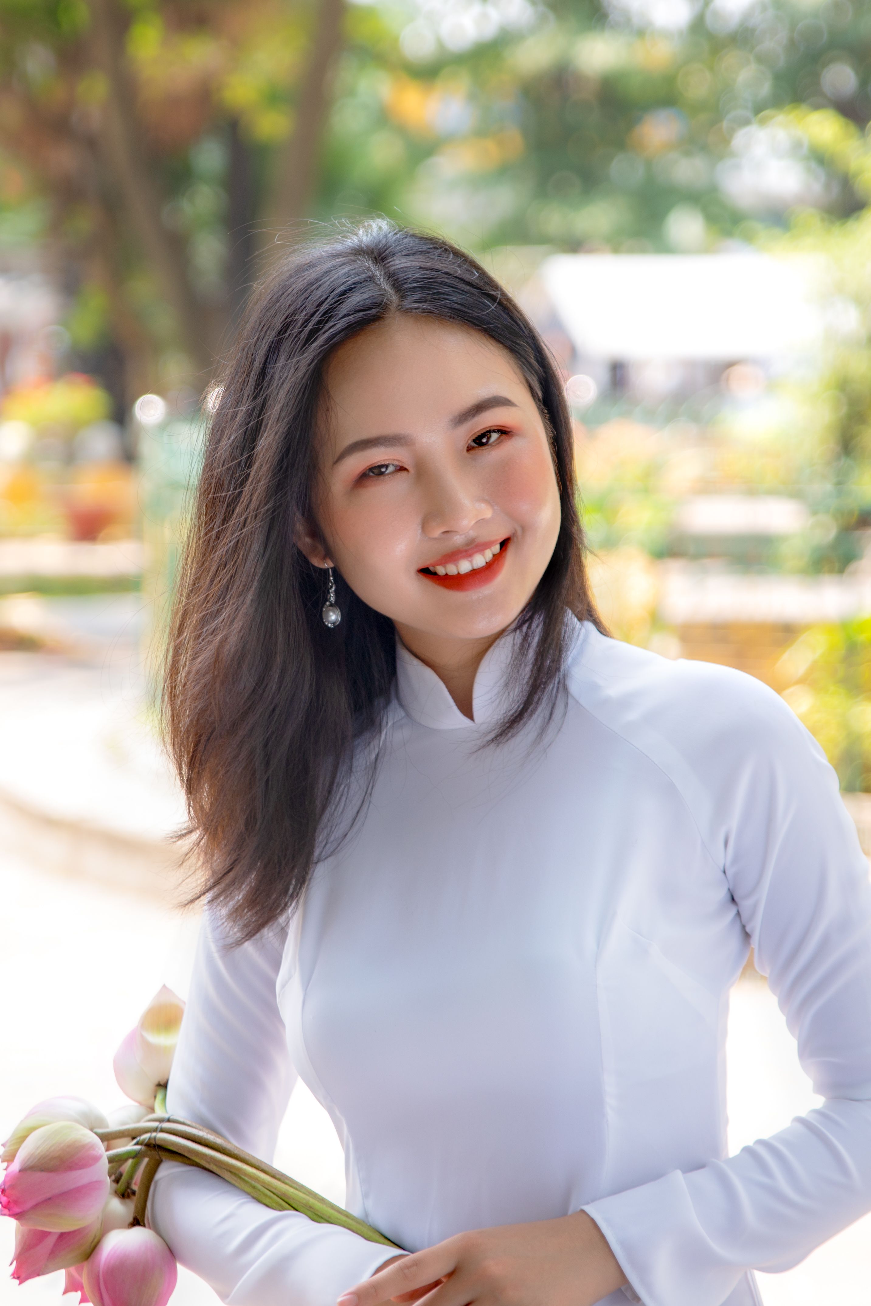 A smiling woman in a white ao dai holding lotus flowers outdoors with a blurred background.