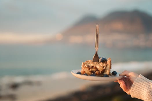 A scenic view of a blueberry pie slice with a fork, held by a hand at a beach.