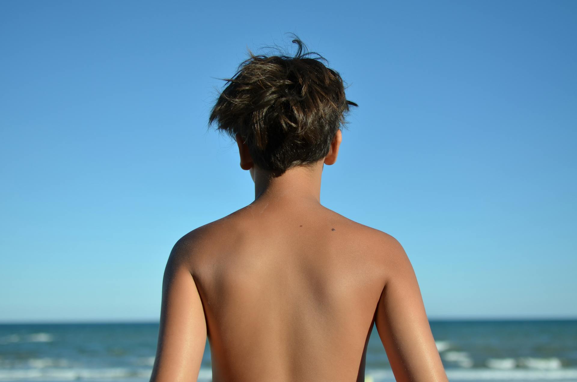 A shirtless child gazes at the ocean with a clear blue sky above.