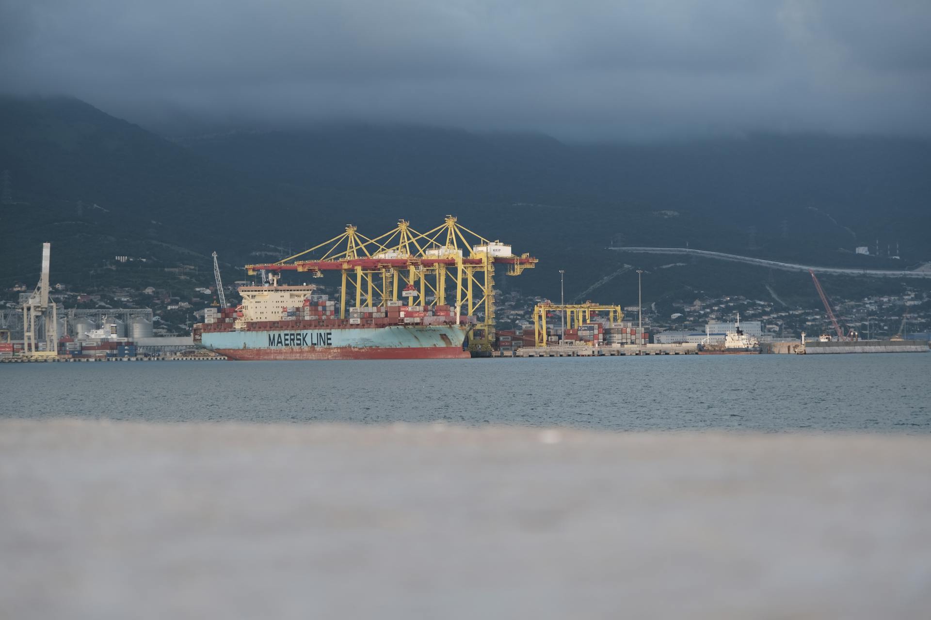 A Maersk cargo ship docked at Novorossiysk port with cranes under cloudy skies.