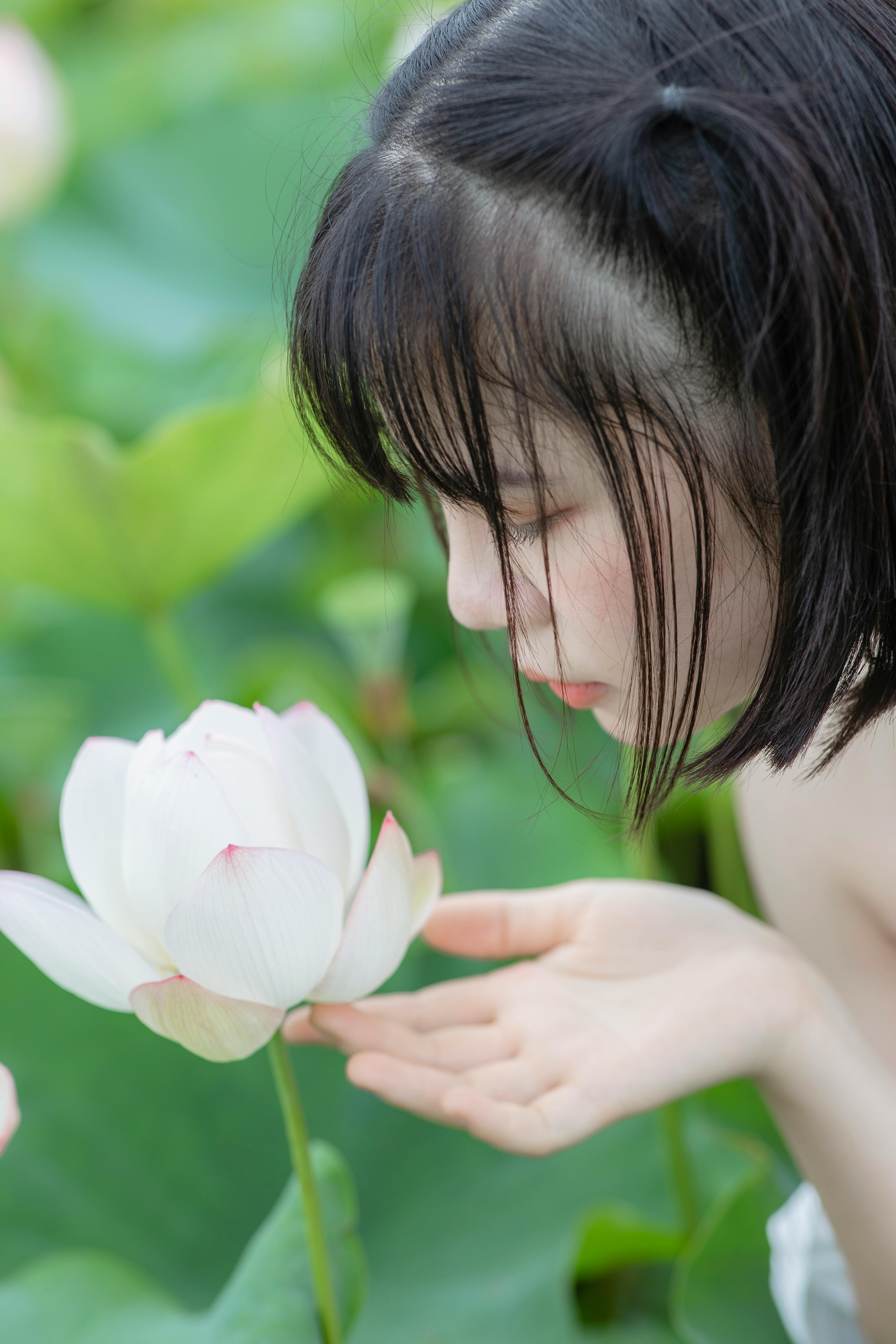 A young woman gently admires a blooming lotus flower in a lush, serene outdoor setting.