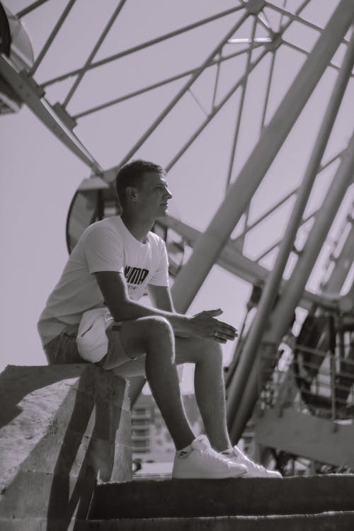 Grayscale Photo of a Man Sitting on Concrete Railing Near a Ferris Wheel