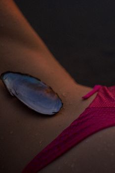 Close-up of a mussel shell resting on skin next to a pink bikini strap. Wet and atmospheric.