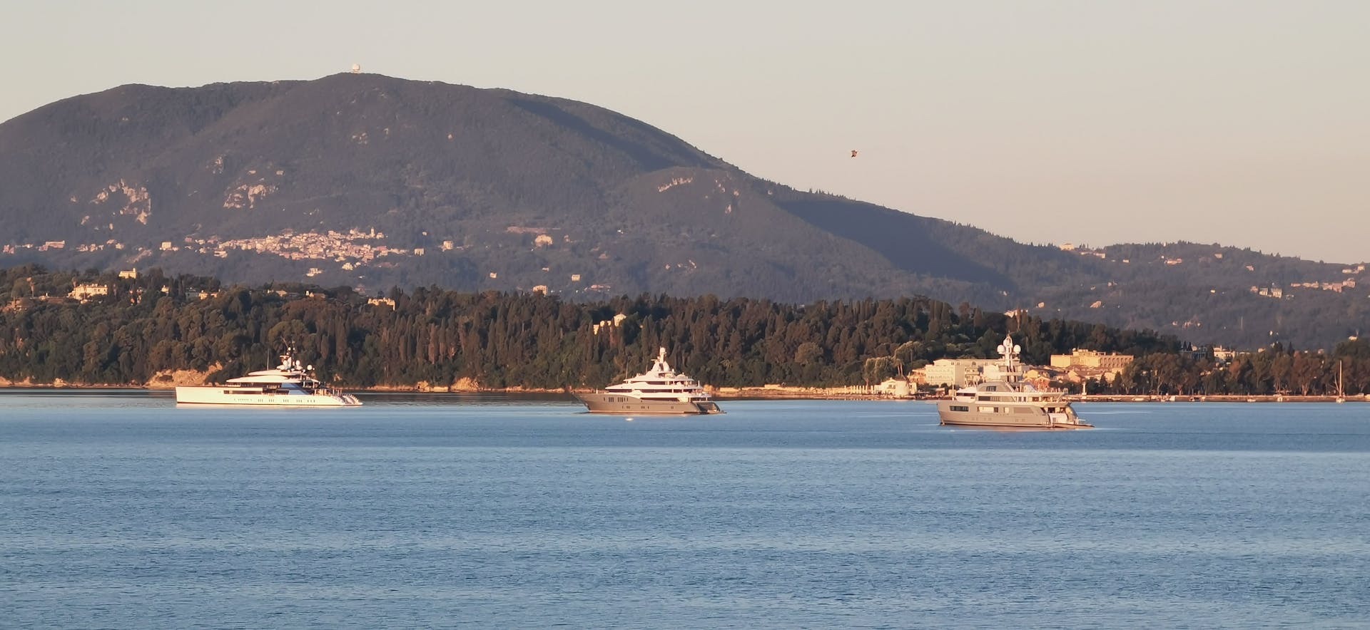 Luxury yachts cruising near Corfu with a mountain backdrop, captured on a sunny day.