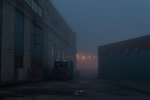 Foggy industrial warehouse yard at night with parked truck and streetlights.