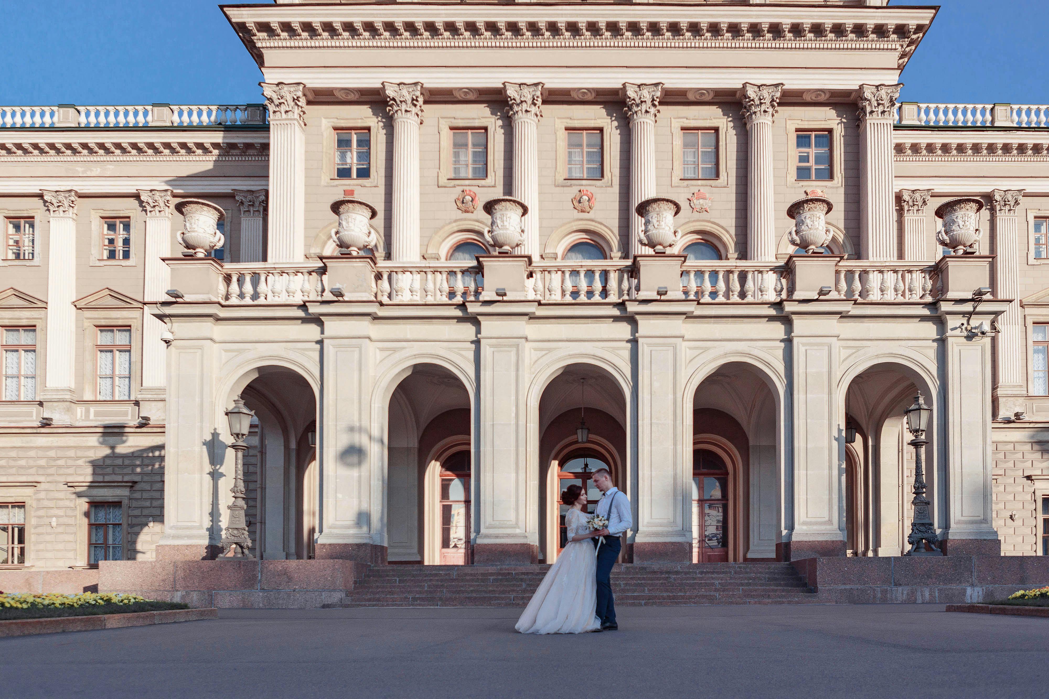 A couple stands in wedding attire in front of the historic Mariinsky Palace in Saint Petersburg.