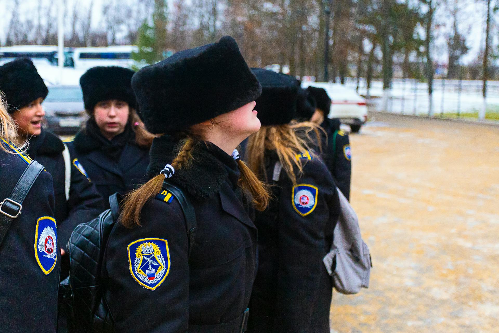 Schoolgirls in matching uniforms and fur hats stand outdoors in a winter setting, displaying lively camaraderie.