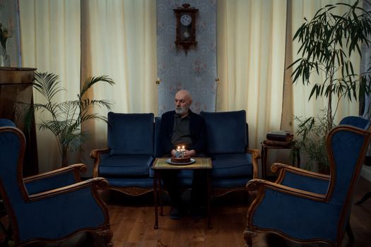 A senior man sitting alone with a birthday cake in an elegant living room setting.