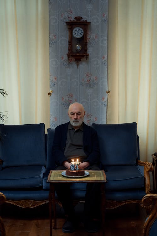 An Elderly Man Sitting on a Couch while Looking at the Cake with Lighted Candles