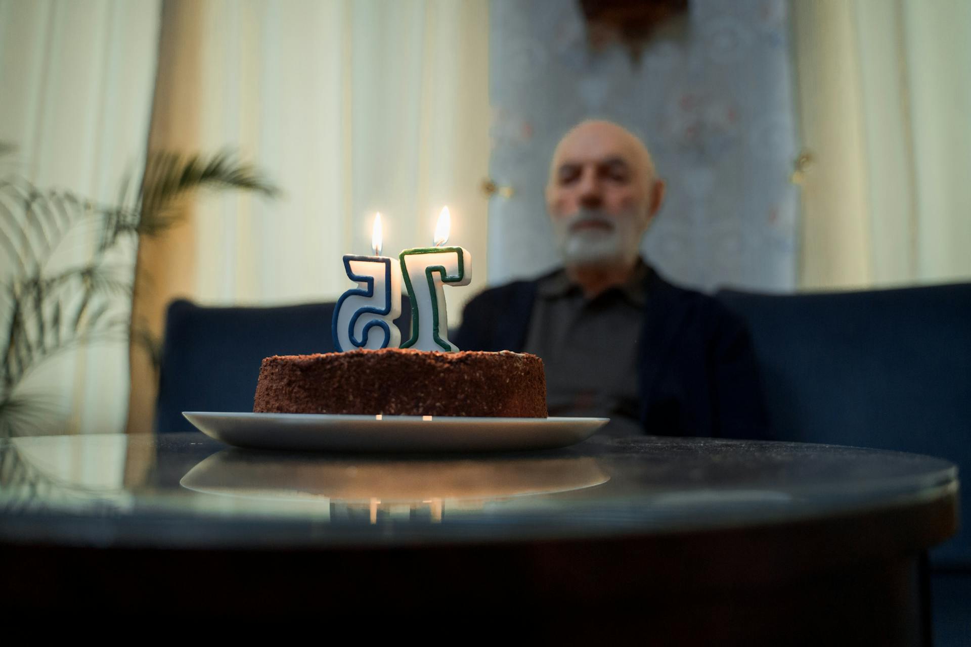 Elderly man sitting alone by a cake with lit candles marking his 75th birthday.