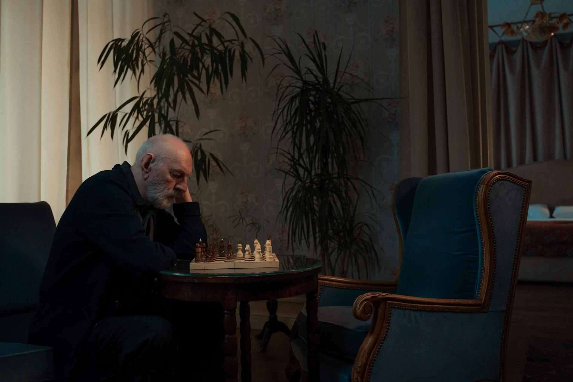 An elderly man sitting alone in a dimly lit room, contemplating a chess move.