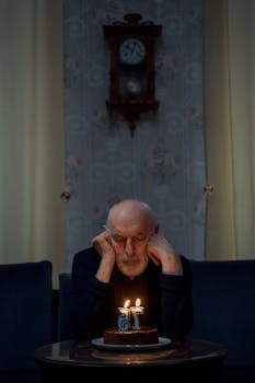 Elderly man reflects quietly by a homemade birthday cake with lit candles in dimly lit room.