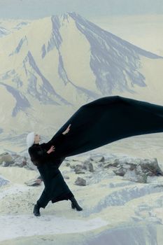 A woman in black cape stands against a snowy mountainous backdrop, creating a dramatic and conceptual winter scene.