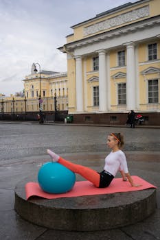 Woman in activewear exercising with a yoga ball on a pink mat outside a historic building.