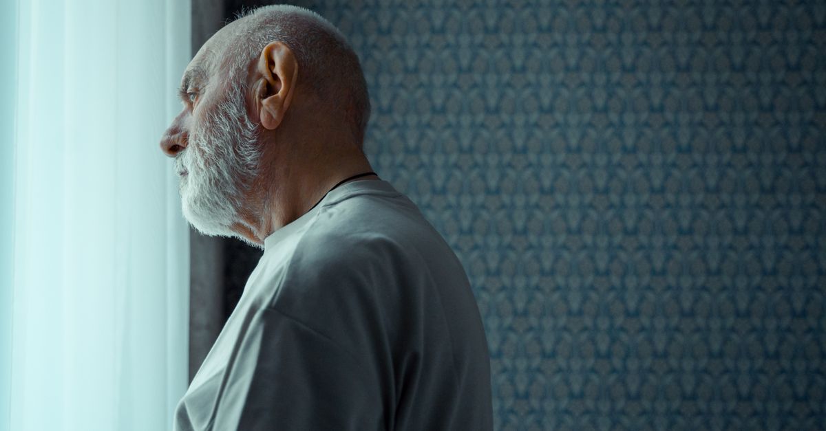 A senior man with a beard looks thoughtfully out of a sunny window, reflecting on life indoors.