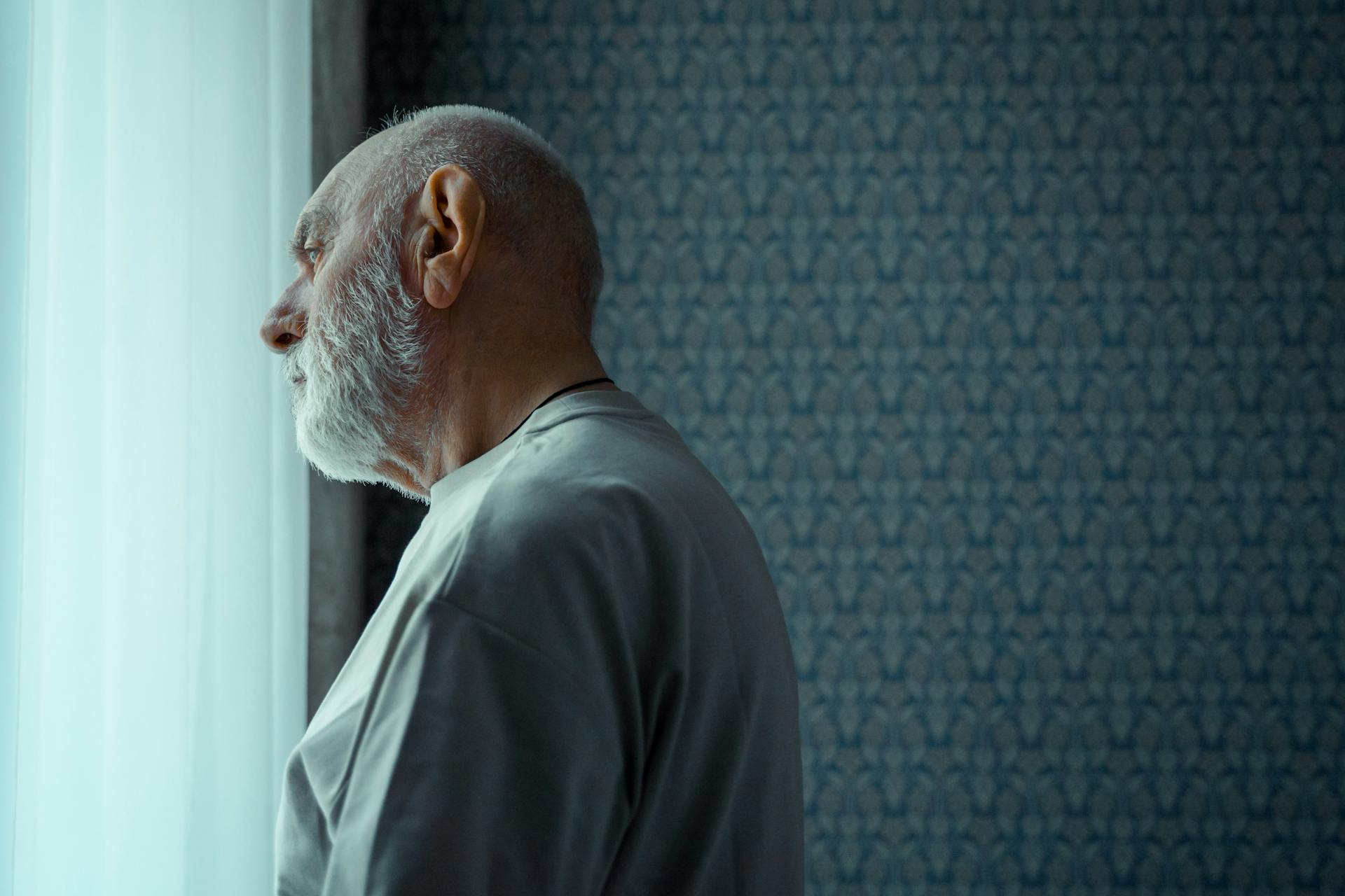 A senior man with a beard looks thoughtfully out of a sunny window, reflecting on life indoors.