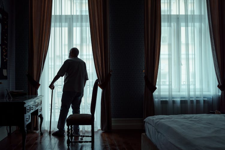 Back View Of An Elderly Man Standing In Front Of The Window