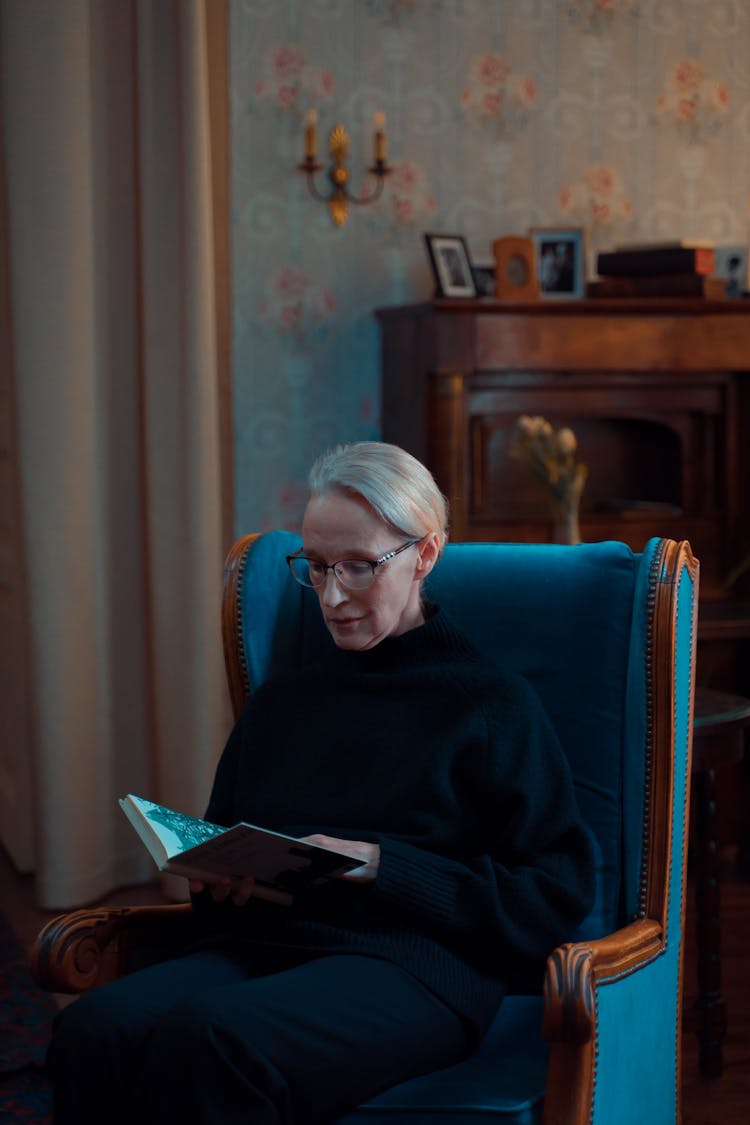 An Elderly Woman In Black Sweater Sitting On The Chair While Reading A Book