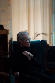 Elegant senior woman with glasses sitting in an armchair inside a cozy room.