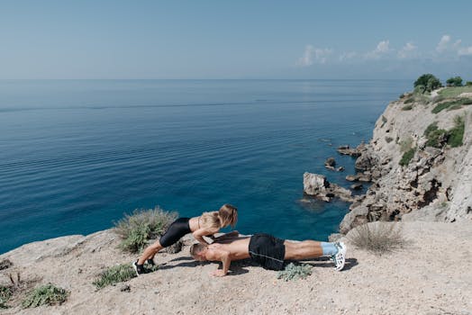 A couple performs a partner workout on a scenic cliffside, overlooking the ocean.