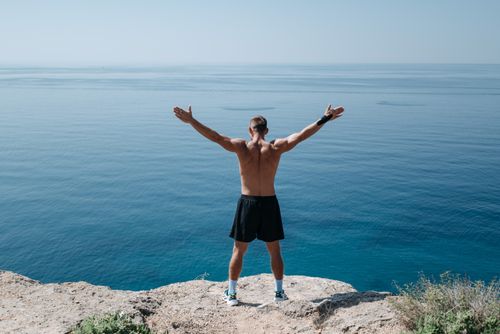 Free A shirtless man stands on a cliff with open arms, embracing freedom and the vast ocean. Stock Photo