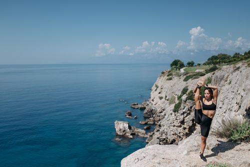 Free Woman in workout attire stretching on a cliff by the sea under a clear blue sky. Stock Photo