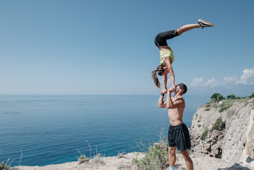 Fit couple performs acrobatic balance pose on a cliffside with ocean view, showcasing strength and flexibility.