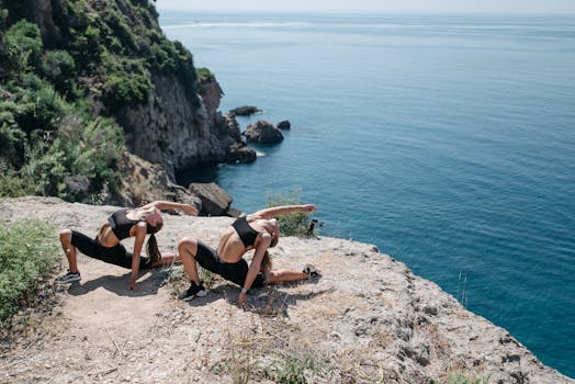Two women in activewear stretching on a coastal cliff with ocean view.