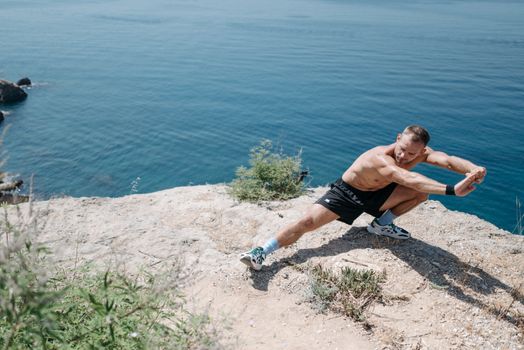 Shirtless man doing a stretch on a cliff overlooking the ocean in daylight.