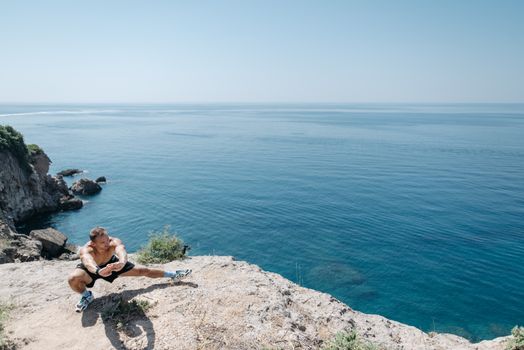 Shirtless man stretching on a cliff's edge by the ocean showcasing a healthy lifestyle and fitness.